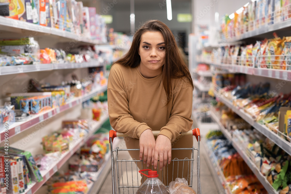 young caucasian lady in a supermarket. Leaning against the cart looking at the camera. Large assortment, complexity of choice. The buyer buys food. young caucasian lady in a supermarket. Leaning against the cart looking at the camera. Large assortment, complexity of choice. The buyer buys food.