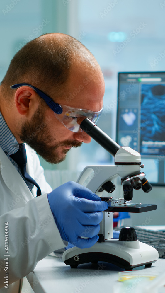 Male scientist looking under microscope in medical development laboratory, entering data in computer. Specialists working on medicine, biotechnology research in advanced pharma lab Male scientist looking under microscope in medical development laboratory, entering data in computer. Specialists working on medicine, biotechnology research in advanced pharma lab