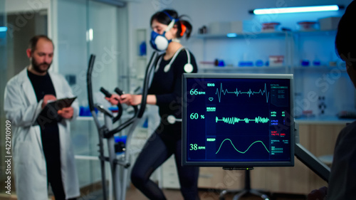 Specialist sport researcher monitoring heart rate on athlete while woman with mask running on cross trainer talking with medical doctor. Physician using tablet controling EKG data in modern laboratory photo