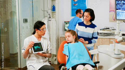 Children sitting on stomatological chair listening pediatric doctor looking on tablet in dental clinic. Dentistry doctor showing to mother of kid teeth radiography using modern gadget in dentist unit