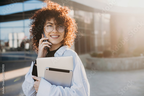 Curly haired businesswoman is posing outside talking on phone while holding a laptop and tablet