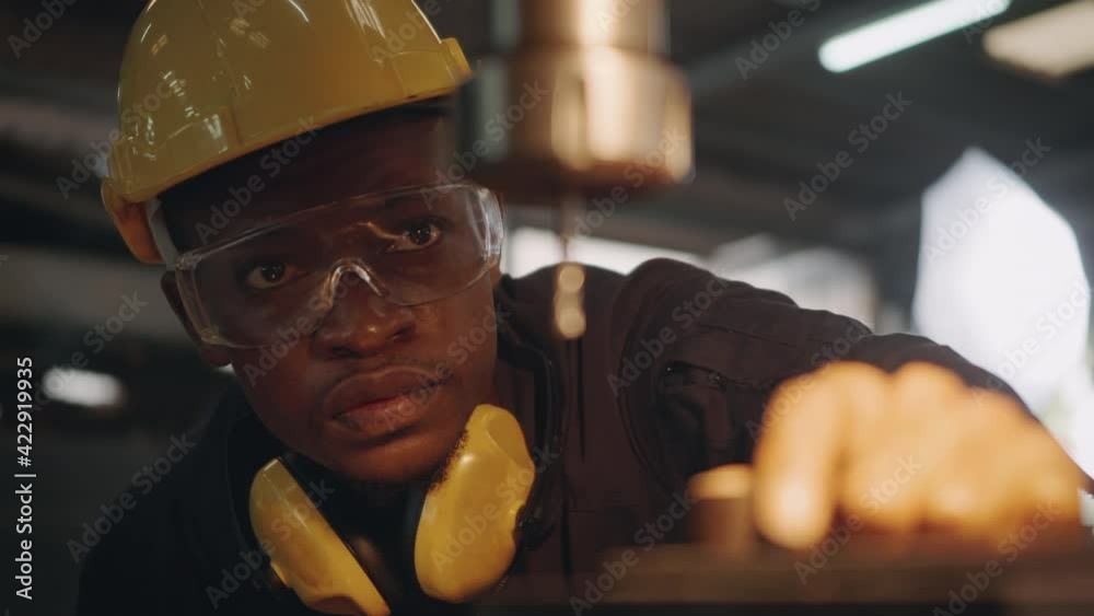 American industrial black young worker man with yellow helmet and ear protection manual rotating on drill machine In order to penetrate workpiece, Engineer at work in industry factory. slow motion