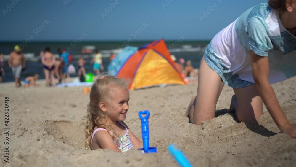Pretty woman bury little girl with sand on beach. Blurred people holidaymakers Pretty woman bury little girl with sand on beach. Blurred people holidaymakers