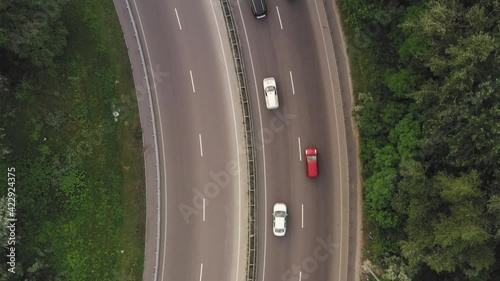 Flying over a curved motorway with traffic: white and black cars and trucks move along the lanes of the highway among the green foliage - aerial overhead shot. Traffic on country road on a summer day.