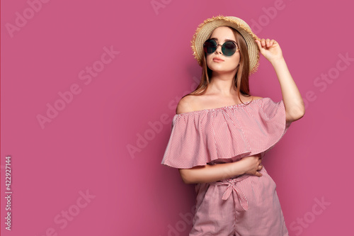 Stylish girl in striped dress standing in straw hat and big round glasses on pink background © zamuruev