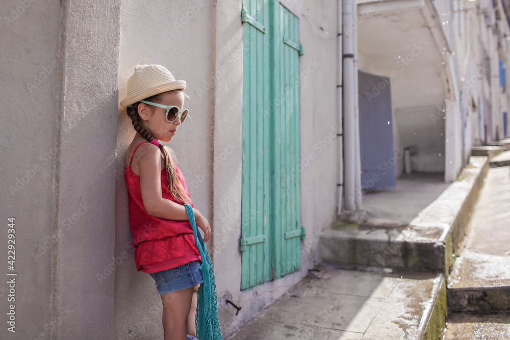 Stylish girl of gen Z in summer hat and mint sunglasses with mesh bag standing near the wooden door at the street of small old town, France, summer family trip, extending vacation with local immersing Stylish girl of gen Z in summer hat and mint sunglasses with mesh bag standing near the wooden door at the street of small old town, France, summer family trip, extending vacation with local immersing