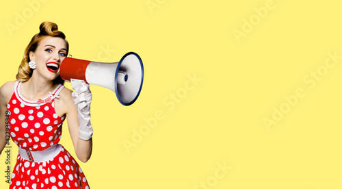 Beauty blond haired woman holding megaphone, shout something. Girl in red pin up style dress in polka dot. Yellow color background. Caucasian model in retro and vintage studio concept.