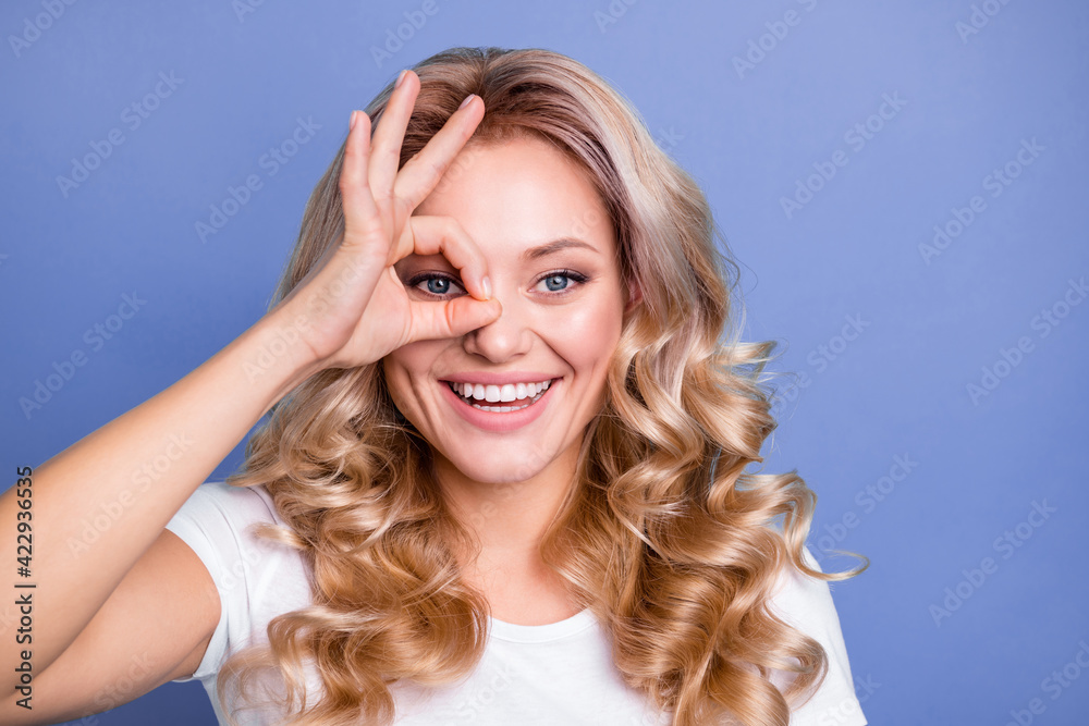 Photo of young happy positive funky funny smiling girl showing ok sing on eye monocular isolated on blue color background Photo of young happy positive funky funny smiling girl showing ok sing on eye monocular isolated on blue color background