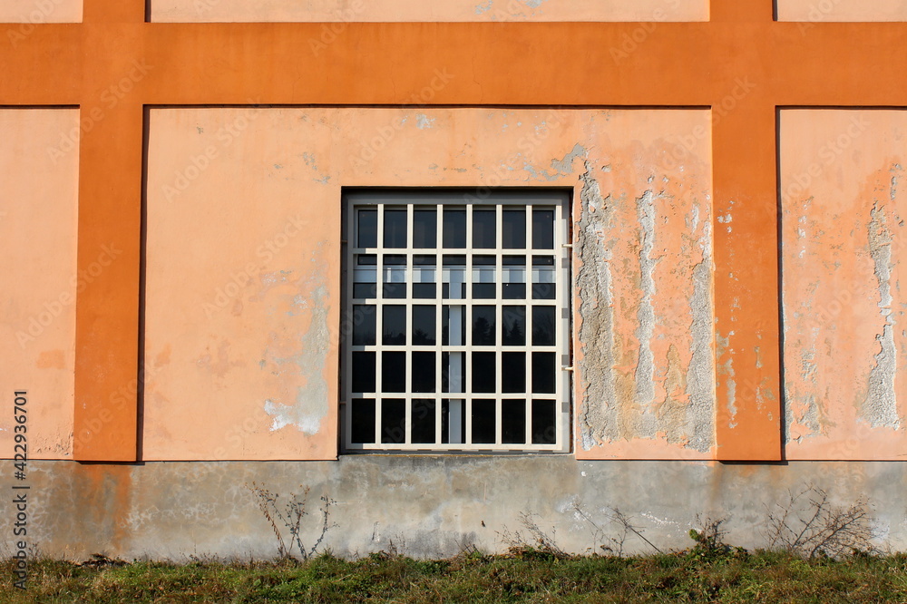 New window with white plastic frame and metal bars protection mounted on colorful old dilapidated industrial factory building wall with cracked facade and uncut grass in front New window with white plastic frame and metal bars protection mounted on colorful old dilapidated industrial factory building wall with cracked facade and uncut grass in front