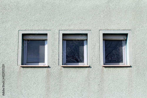 Three small windows in a row with tree branches in reflection and white plastic frame covered with dust and cobwebs on top of closed dirty blinds boxes mounted on light green family house wall