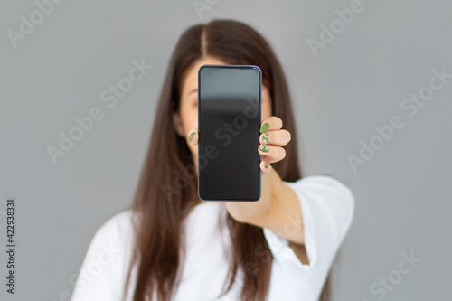 Close up portrait of a smiling woman showing blank screen mobile phone while standing isolated over gray background
