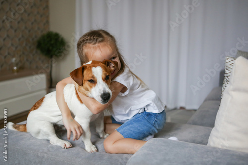 Happy child with a dog. Portrait of a girl with a pet. The child plays and hugs the puppy. Little girl and a puppy on the couch. Pet at home. Taking care of animals. © Valeriya