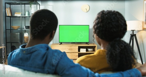 Close up of wife and husband sitting at home together watching TV with chroma key. Back view of African American couple man and woman sitting on sofa watch television program, green screen