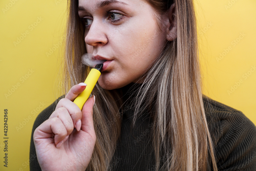 A young girl smokes disposable electronic cigarette. Bright yellow background A young girl smokes disposable electronic cigarette. Bright yellow background