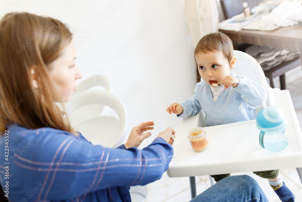 Mother feed little toddler baby boy with open mouth from plastic spoon in high chair view from back Mother feed little toddler baby boy with open mouth from plastic spoon in high chair view from back