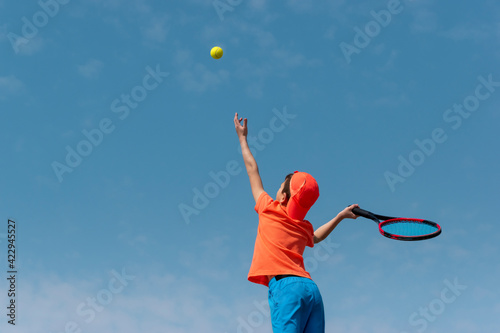 Child boy tennis player in orange sportswear with racket learning to performs serve ball. Kids sport tennis game, training at school or club. Child athlete in action. Background copy space © Elena