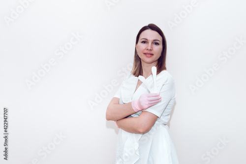 Doctor beautician in a white medical gown on a gray background holds a cosmetic spatula with place for text or logo. A woman in a white medical gown holds a white plastic cosmetic spatula.