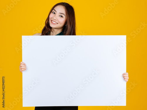 Cute and pretty curly hair Asian female brunette holding white blank board poses to camera with a joyful for advertising and banner use purpose, studio shot isolated on bright yellow background