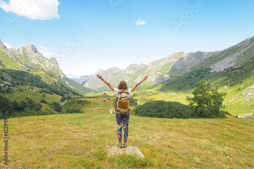 A girl with a backpack and arms wide open in the air is looking to the mountains with a blue sky and white clouds.