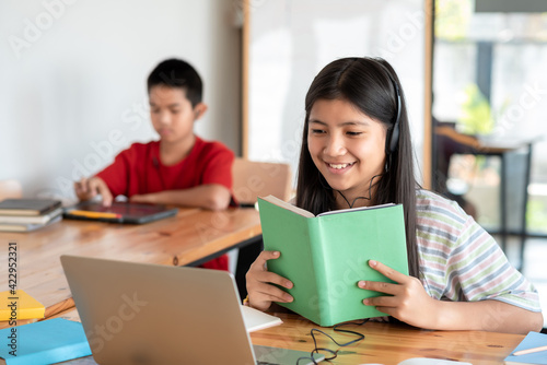 Image of a happy Asian girl wearing headphones holding a textbook online learning using a laptop in the classroom, the background is blurred.