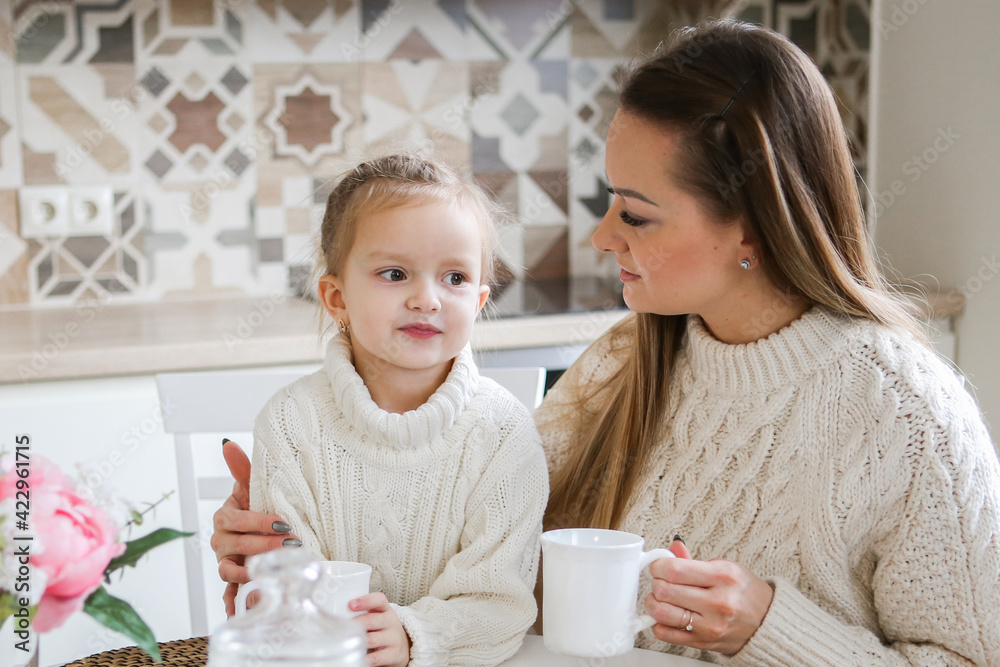 Loving young mother in a sweater is talking to a cute funny baby daughter enjoying time together at home in the kitchen with tea. happy family. mom with little baby girl fun play feel joy hugging Loving young mother in a sweater is talking to a cute funny baby daughter enjoying time together at home in the kitchen with tea. happy family. mom with little baby girl fun play feel joy hugging
