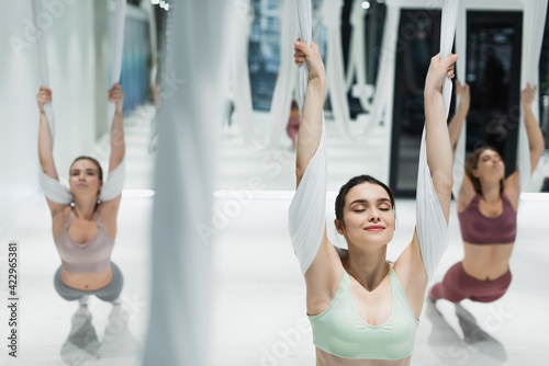 group of young sportswomen stretching with aerial yoga straps on blurred foreground