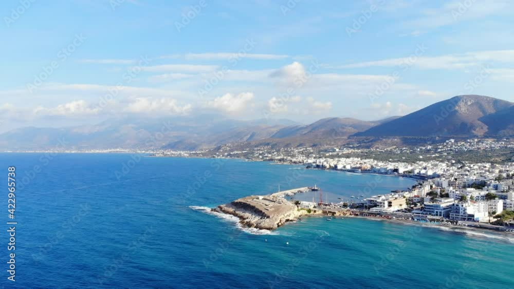 Scenic panorama of North Crete at mellow season, half-island at Hersonissos port, many buildings on land. Tall mountains seen on background. Crystal clear and transparent water of Aegean sea Scenic panorama of North Crete at mellow season, half-island at Hersonissos port, many buildings on land. Tall mountains seen on background. Crystal clear and transparent water of Aegean sea
