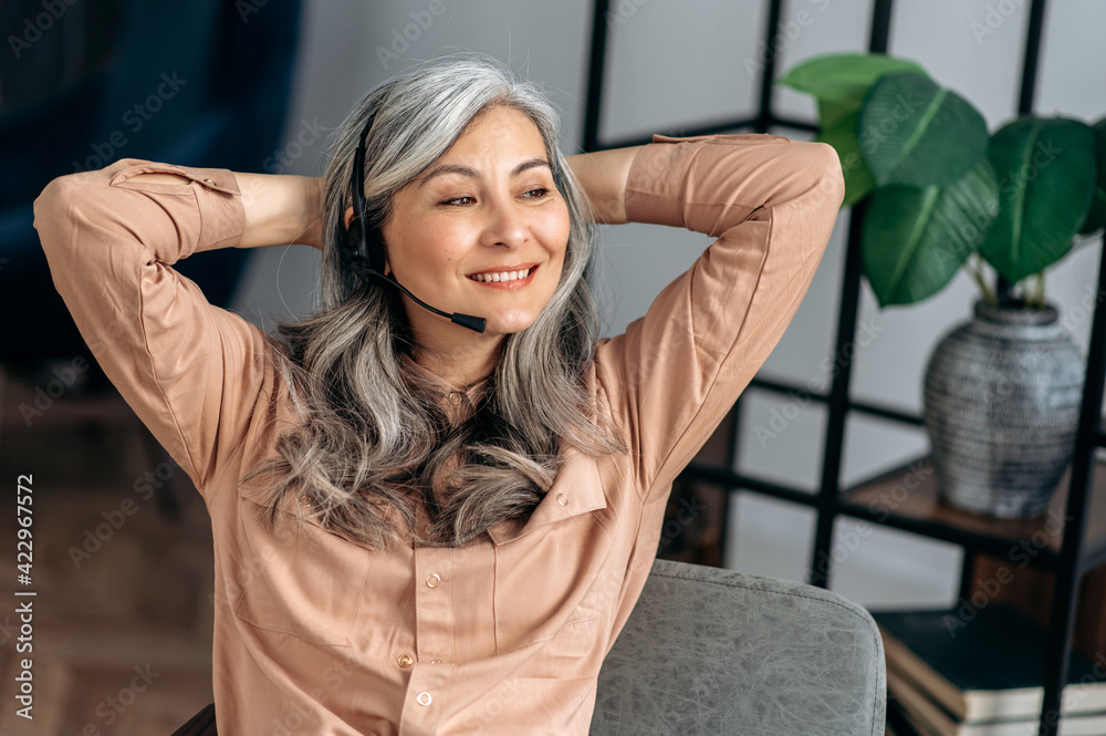 Joyful successful influential senior gray-haired asian business woman, top manager, freelancer, sitting at the office, taking a break during work, crossing her arms behind her head, smiling