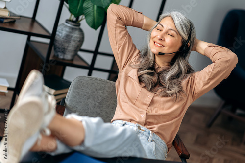 Relaxed beautiful mature gray-haired asian business woman, freelancer, sitting in the office, throwing her legs on table, taking break during work, crossing her arms behind her head, smiling © Kateryna