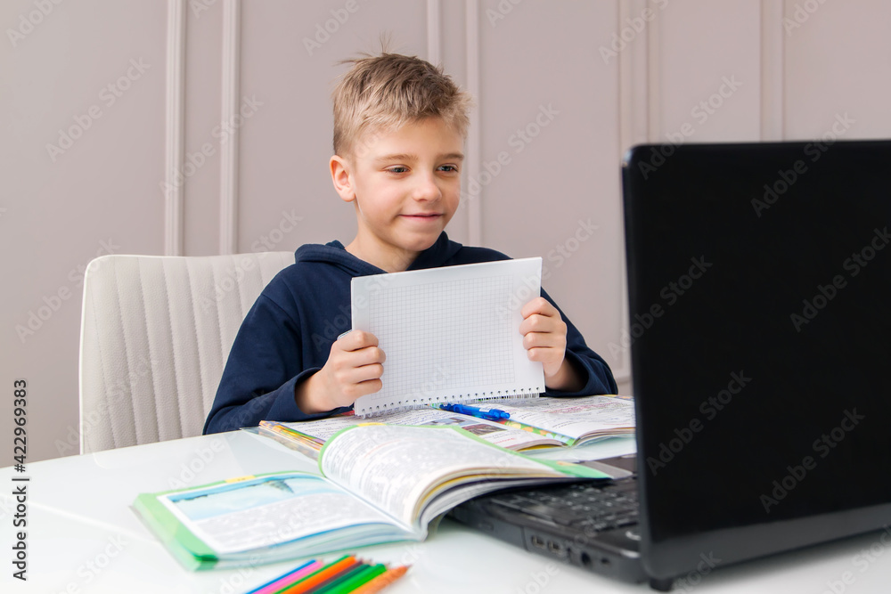 Boy shows notebook with answer to teacher on laptop screen. Smart Little Boy Is doing His Homework in Living Room. COVID-19 pandemic forces children online learning Boy shows notebook with answer to teacher on laptop screen. Smart Little Boy Is doing His Homework in Living Room. COVID-19 pandemic forces children online learning