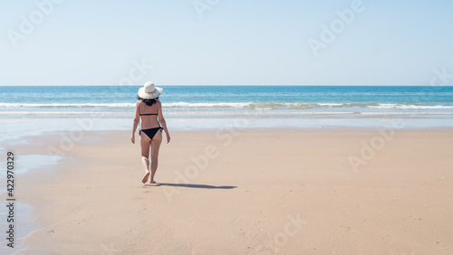 Young brunette woman with curly hair wearing black bikini and hat walking along a lonely beach. vacation, leisure, relaxation and tranquility concept