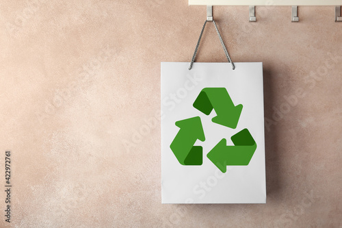Paper shopping bag with recycling symbol hanging on beige wall. Space for text photo
