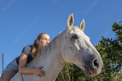 junge blonde Frau liegt vertrauensvoll auf dem Hals ihres Schimmels und schaut vor blauem Himmel in die Sonne photo