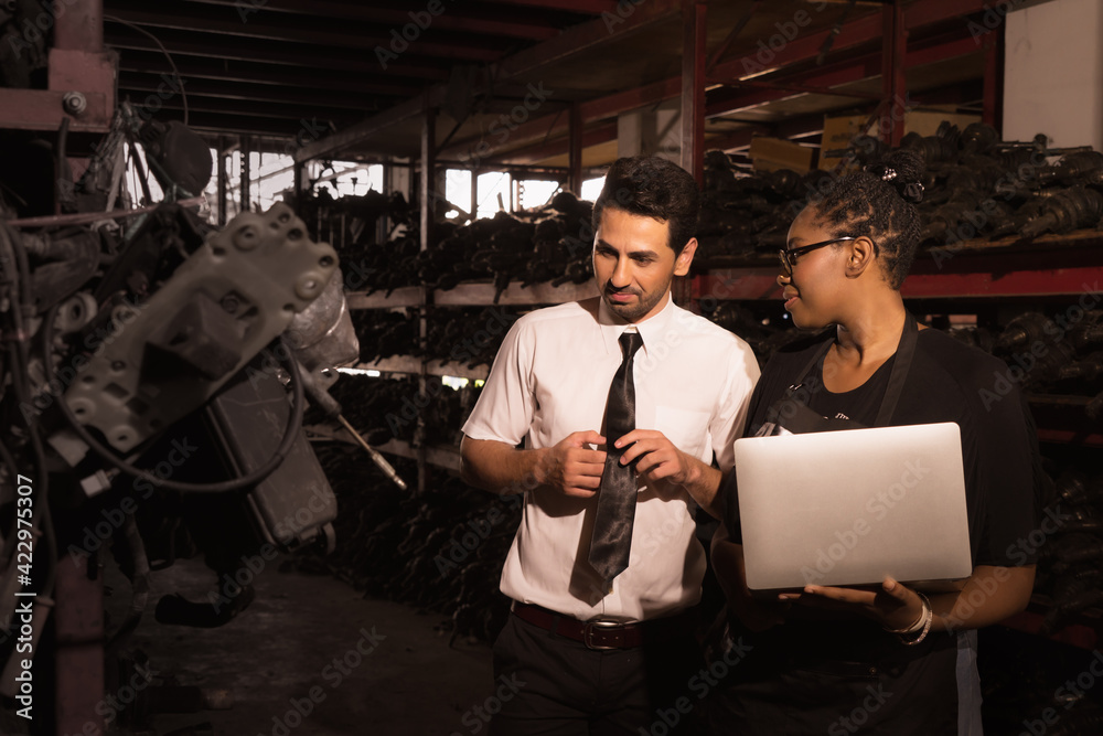 Diversity of two people, a caucasian business manager looking at car part in factory-warehouse and serious talking with black African worker woman who holding laptop-notebook