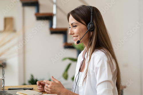 Happy employee woman with headphones looking at laptop computer screen, helping customers remotely, talking in online video call, working from home due to lockdown, pandemic covid-19. Distance job.
