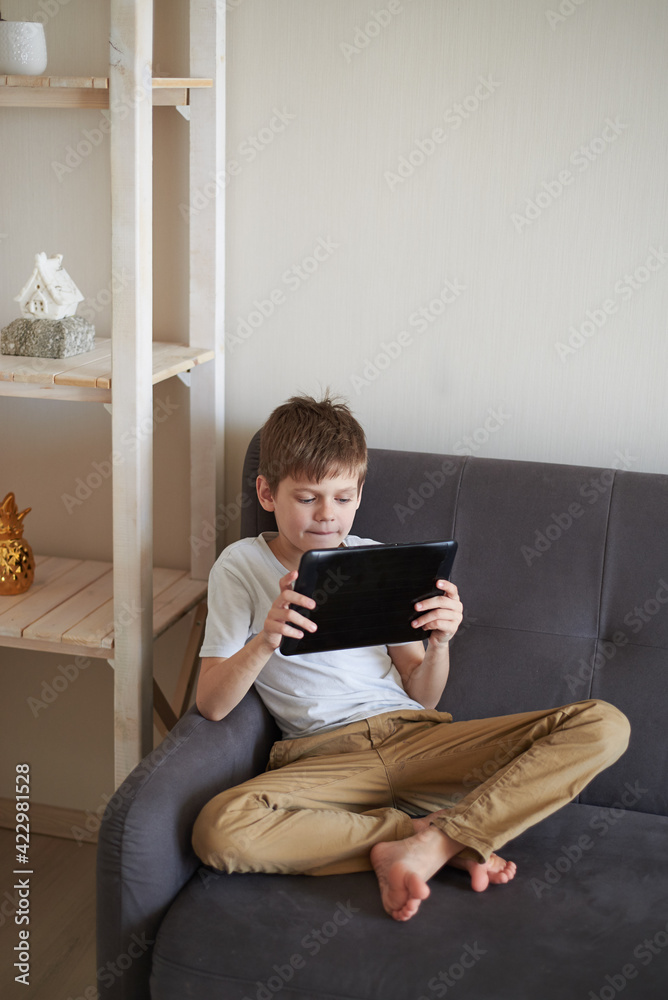 The boy sits at home on the sofa and holds a tablet in his hands