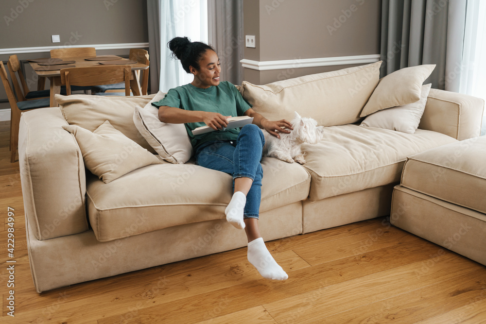 Happy black woman stroking her dog while reading book on sofa at home