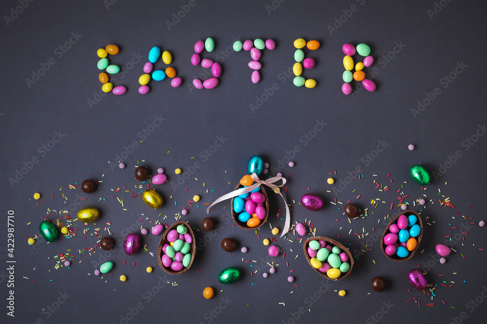 Chocolate Easter eggs full of candies, surrounded by colorful sweets on the grey background. Easter background. Easter sign made with candies