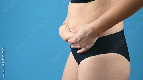 Cropped view of woman in black panties holds her belly fat on blue background. Body control, diet and weight loss concept photo