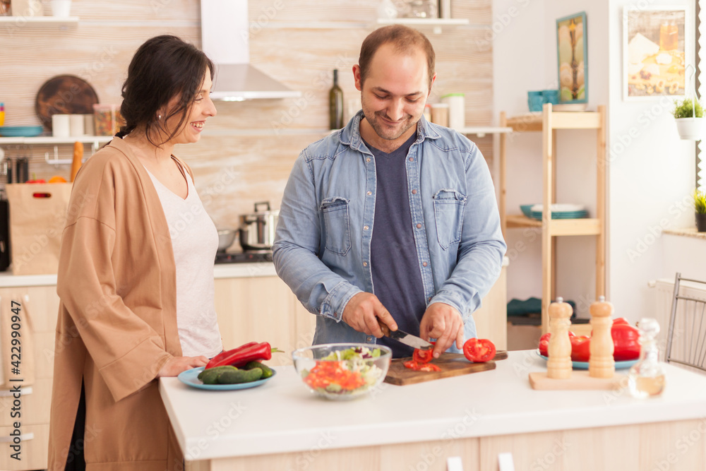 Man slicing tomatoes for salad on cutting board in kitchen while wife is smiling. Happy in love cheerful and carefree couple helping each other to prepare meal Man slicing tomatoes for salad on cutting board in kitchen while wife is smiling. Happy in love cheerful and carefree couple helping each other to prepare meal