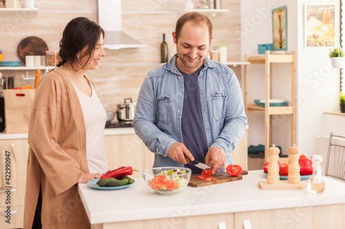 Man slicing tomatoes for salad on cutting board in kitchen while wife is smiling. Happy in love cheerful and carefree couple helping each other to prepare meal