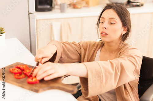 Woman with walking handicap cutting red paper on wooden board in kitchen using sharp knife. Disabled paralyzed handicapped woman with walking disability integrating after an accident. photo