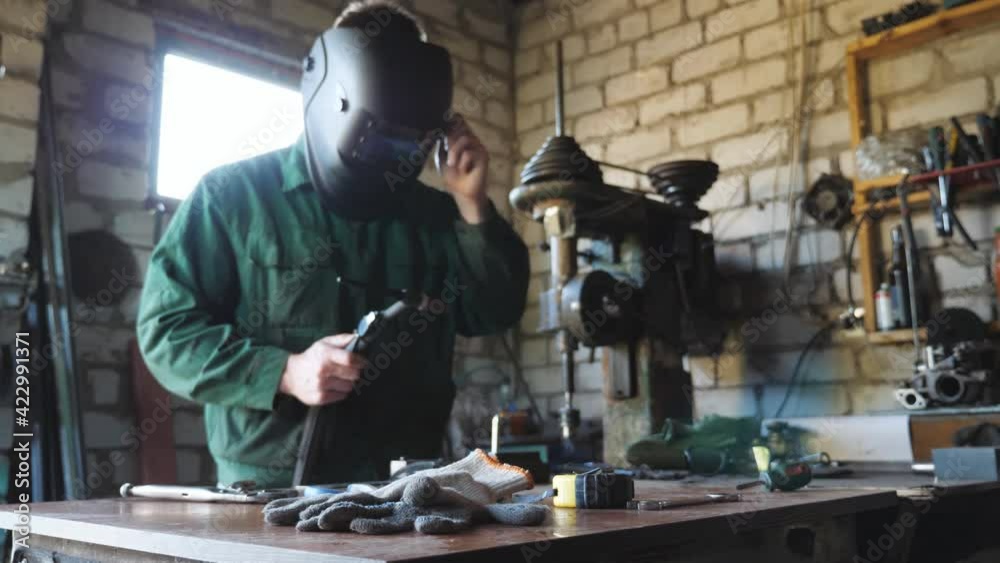 Young mechanic in protective mask holding broken welding machine and trying to solder some metal details at garage. Male welder working at his workshop. Concept of maintenance service. Close up