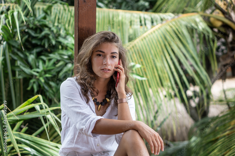 Portrait of young beautiful woman freelancer with red smartphone on balcony of tropical bungalow with palm trees view Portrait of young beautiful woman freelancer with red smartphone on balcony of tropical bungalow with palm trees view