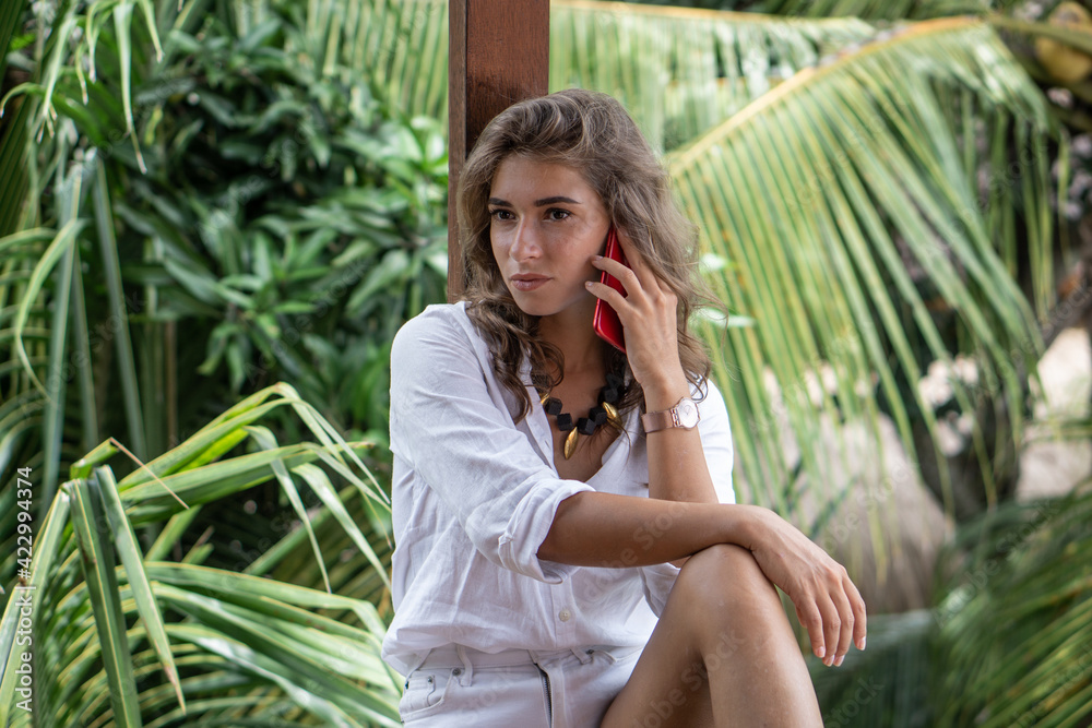 Portrait of young beautiful woman freelancer with red smartphone on balcony of tropical bungalow with palm trees view Portrait of young beautiful woman freelancer with red smartphone on balcony of tropical bungalow with palm trees view