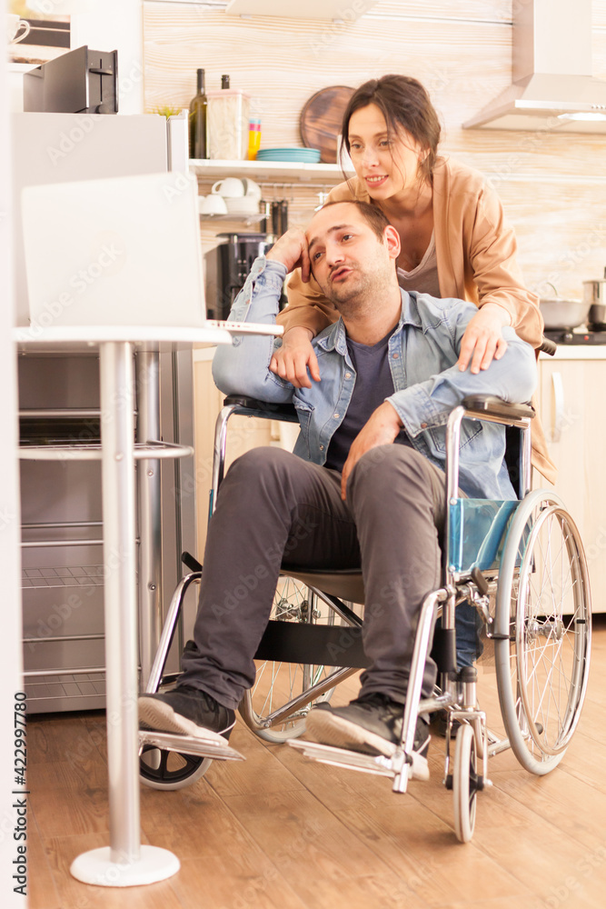 Fototapeta premium Handicapped man in wheelchair and wife during video conference on laptop in kitchen. Disabled paralyzed handicapped man with walking disability integrating after an accident.