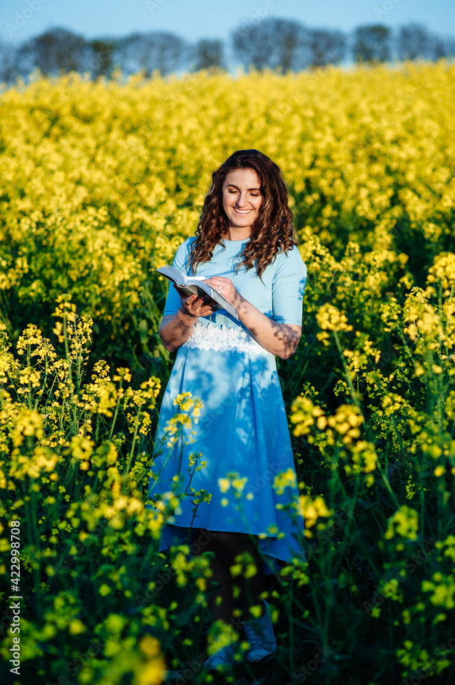 Portrait of a happy female student holding books and looking at camera outdoors. Young, beautiful girl holding a book, on a background of yellow flowers in a field Portrait of a happy female student holding books and looking at camera outdoors. Young, beautiful girl holding a book, on a background of yellow flowers in a field