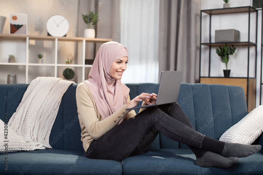 Working and learning remotely, freelance concept. Modern confident 30-aged arab girl in hijab using laptop at home, typing while sitting on soft blue sofa in living room. Working and learning remotely, freelance concept. Modern confident 30-aged arab girl in hijab using laptop at home, typing while sitting on soft blue sofa in living room.