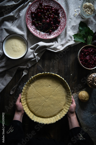 Step by step process of making cherry custard pie. Woman holding baking form with dough on table with custard, cherries and almond flakes on it.