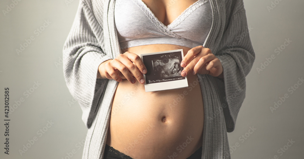 Pregnant woman holding an ultrasound picture next to her pregnant belly. The first photo of the baby. Maternal prenatal care. Maternity, pregnancy, baby expectation concept. Copy space Pregnant woman holding an ultrasound picture next to her pregnant belly. The first photo of the baby. Maternal prenatal care. Maternity, pregnancy, baby expectation concept. Copy space